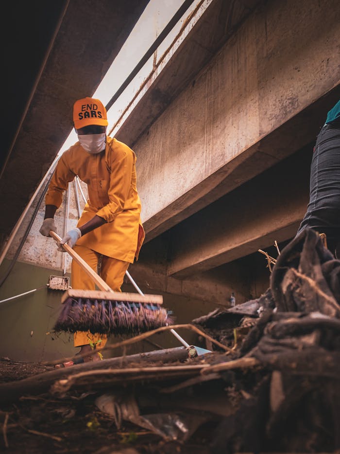 Man in uniform sweeping trash under a concrete bridge, promoting cleanliness and awareness.